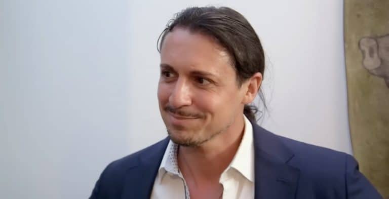 A man with long brown hair in a ponytail. He's wearing a white shirt under a blue suit jacket.